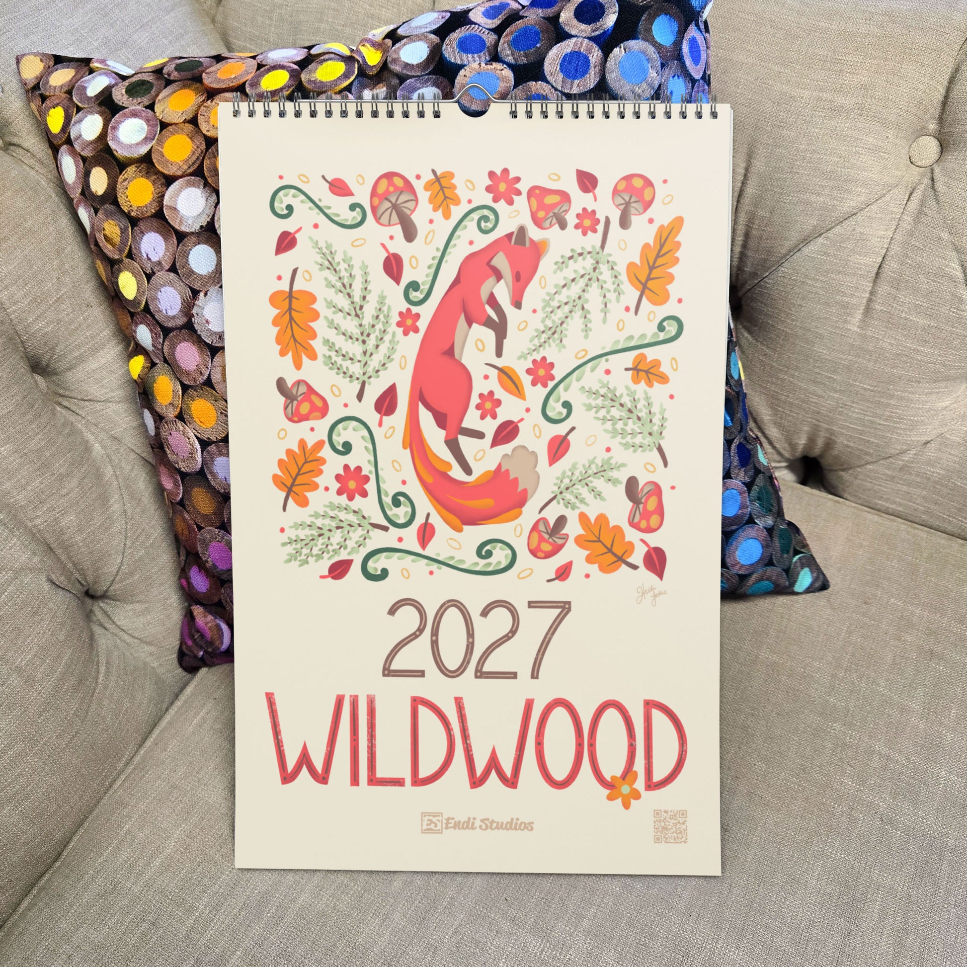 A 2027 spiral-bound wall calendar titled 'WILDWOOD' by  Endi Studios, featuring a colorful illustration of a fox and autumn leaves, styled on a beige tufted sofa with patterned cushions.