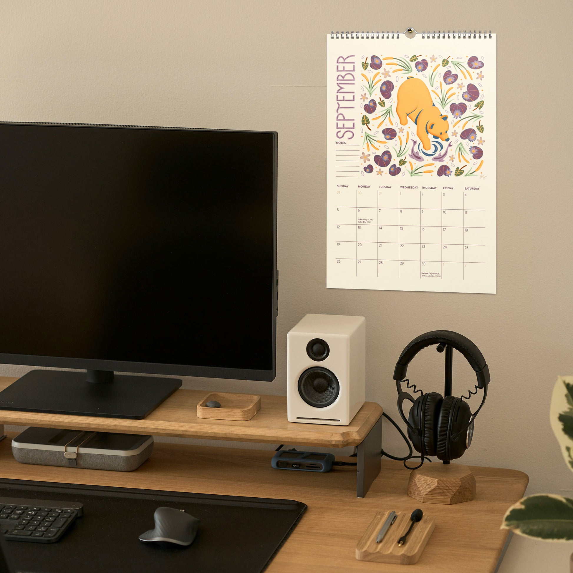 A September wall calendar with a colorful illustration of a bear and flowers hangs on a beige wall above a wooden desk setup with a monitor, speaker, headphones, and other desk accessories. 2027 wire-bound wall calendar hanging on a neutral wall, displaying colourful folk-art animal artwork and a clear monthly layout. Ideal for home offices, kitchens, studios, classrooms, or anyone looking for a decorative illustrated 2027 wall calendar with unique art.