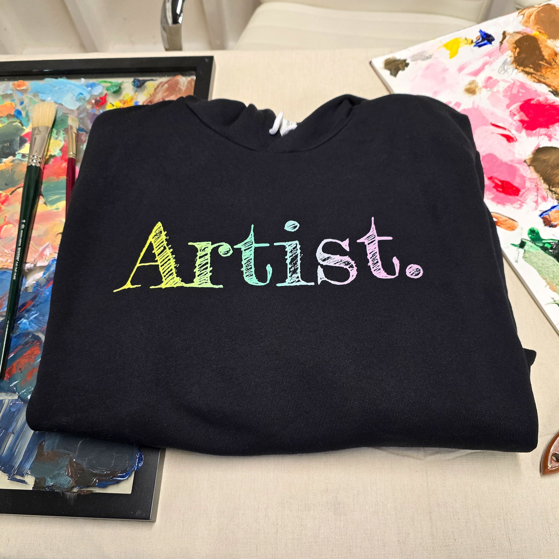 Close-up of the hoodie on an artist's background. A black artist hoodie is displayed in a flat lay. The hoodie features a sketch-style hand-drawn gradient text design in yellow, green and light purple, ideal for artists, creatives, students and illustrators. This aesthetic art hoodie is perfect for year-round outfits and makes a thoughtful gift for art lovers.