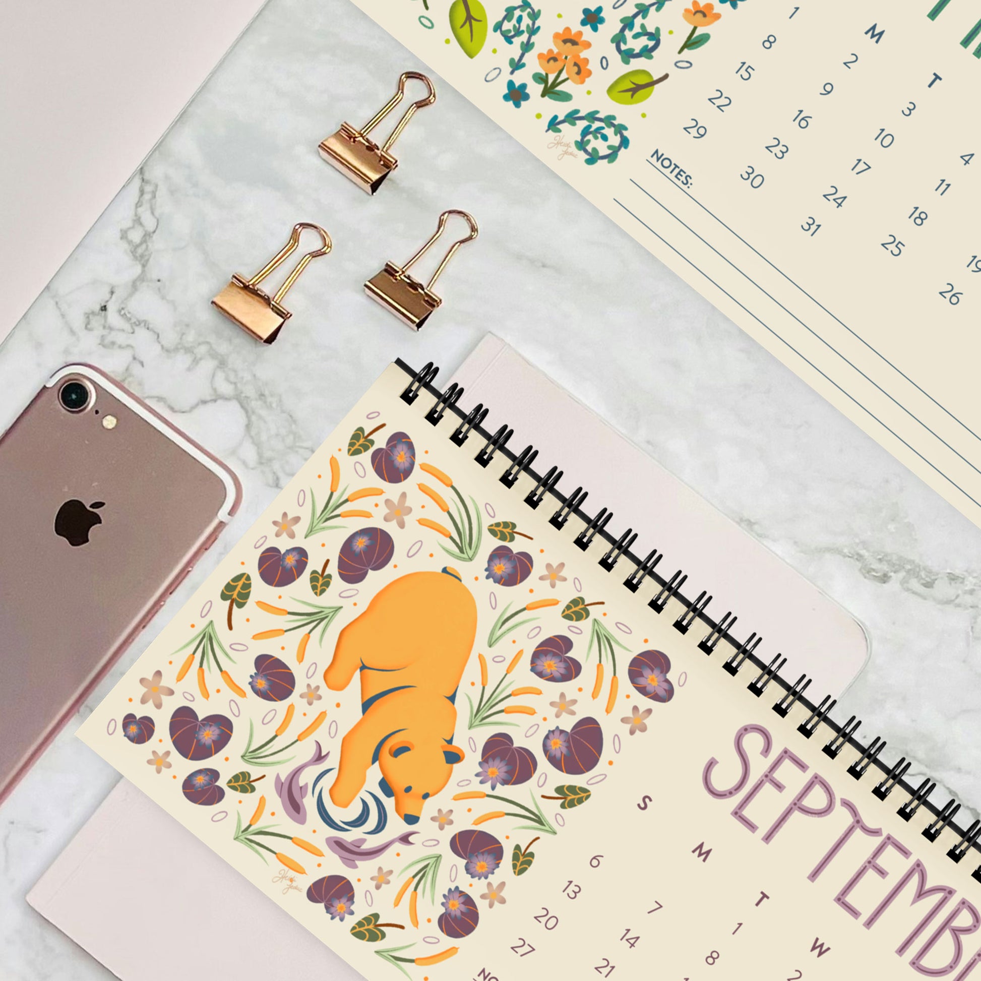A flat-lay photo of the 2026 desk calendar shown open on a clean desktop surface. The satin-finish pages display bright folk-art animal illustrations with a clear monthly layout. Black spiral binding and the tent-card base are partially visible, highlighting the calendar’s compact, freestanding design. Perfect for shoppers wanting a small 2026 desk calendar for office décor, work-from-home setups, or cute illustrated organizational tools.