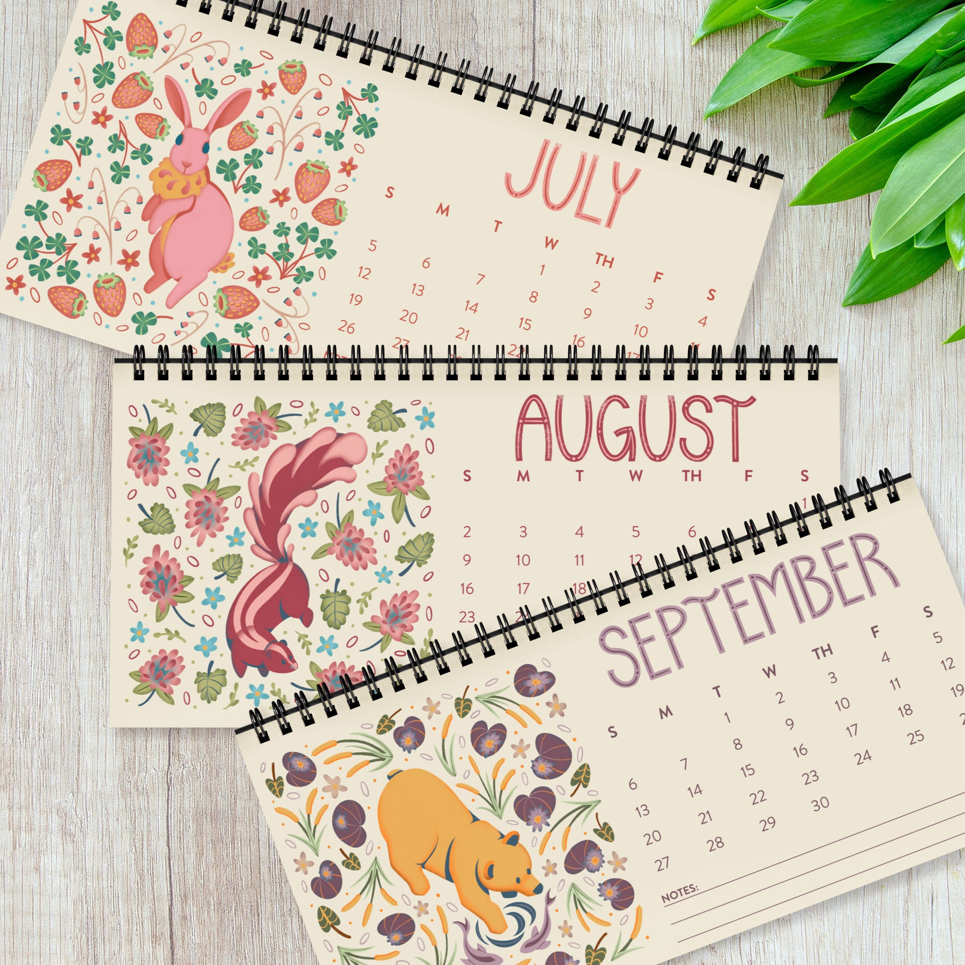 Three months from the 2026 desk calendar displayed together, each featuring a unique folk-art animal illustration printed on smooth satin paper. The monthly pages show vibrant artwork, clear date grids, and black spiral binding at the top. This set of quarterly pages highlights the high-quality printing and cohesive visual style of the calendar, ideal for buyers looking for a colourful, small standing desk calendar with illustrated animal art for 2026.