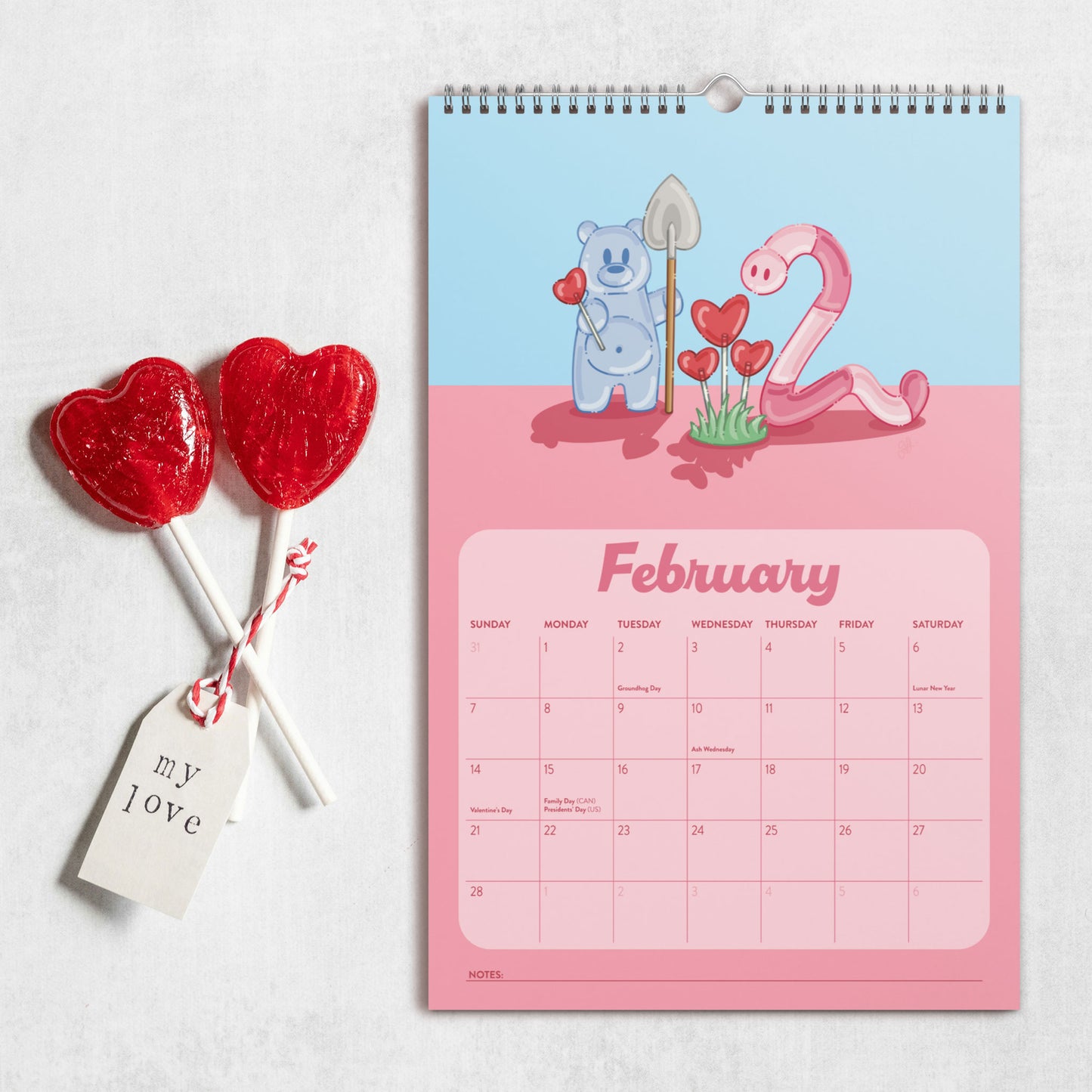 A spiral-bound February 2027 wall calendar with a Valentine's Day theme, featuring a light blue top half and pink bottom half, styled next to two red heart-shaped lollipops on a light gray textured background.
