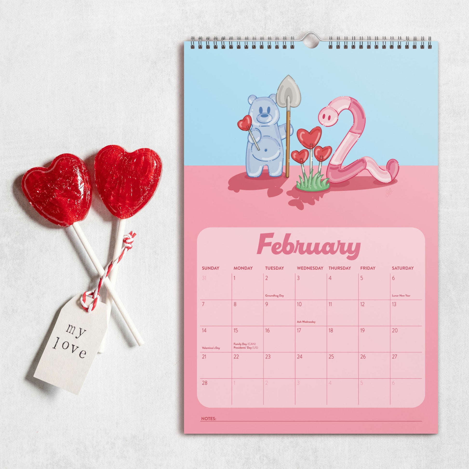 A spiral-bound February 2027 wall calendar with a Valentine's Day theme, featuring a light blue top half and pink bottom half, styled next to two red heart-shaped lollipops on a light gray textured background.