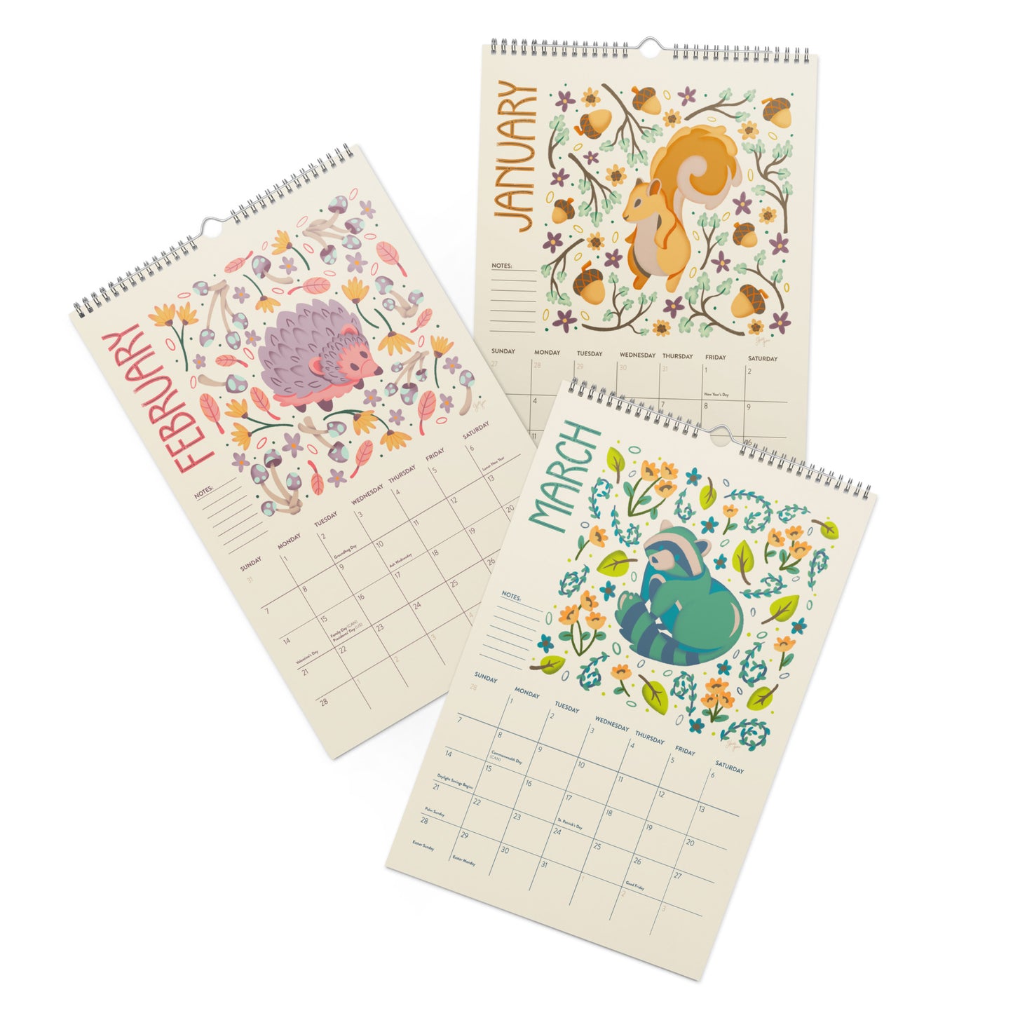 Three spiral-bound monthly wall calendars fanned out on a white background, each featuring a different spring animal illustration: a purple hedgehog in February, an yellow squirrel in January, and a green racoon in March, surrounded by floral and acorn motifs. A 2027 wall calendar featuring colourful folk-art animal illustrations displayed in a flat lay. Wire-bound with premium thick paper, this illustrated monthly calendar showcases vibrant artwork for each month. Ideal for shoppers looking for a 2027 gift