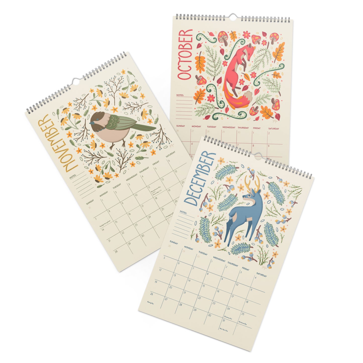 Three spiral-bound wall calendars fanned out on a white background, each showing a different month with a colorful nature illustration: a bird in November, a fox in October, and a deer in December. A 2027 wall calendar featuring colourful folk-art animal illustrations displayed in a flat lay. Wire-bound with premium thick paper, this illustrated monthly calendar showcases vibrant artwork for each month. Ideal for shoppers looking for a 2027 calendar, animal art calendar, home décor calendar, or unique artis