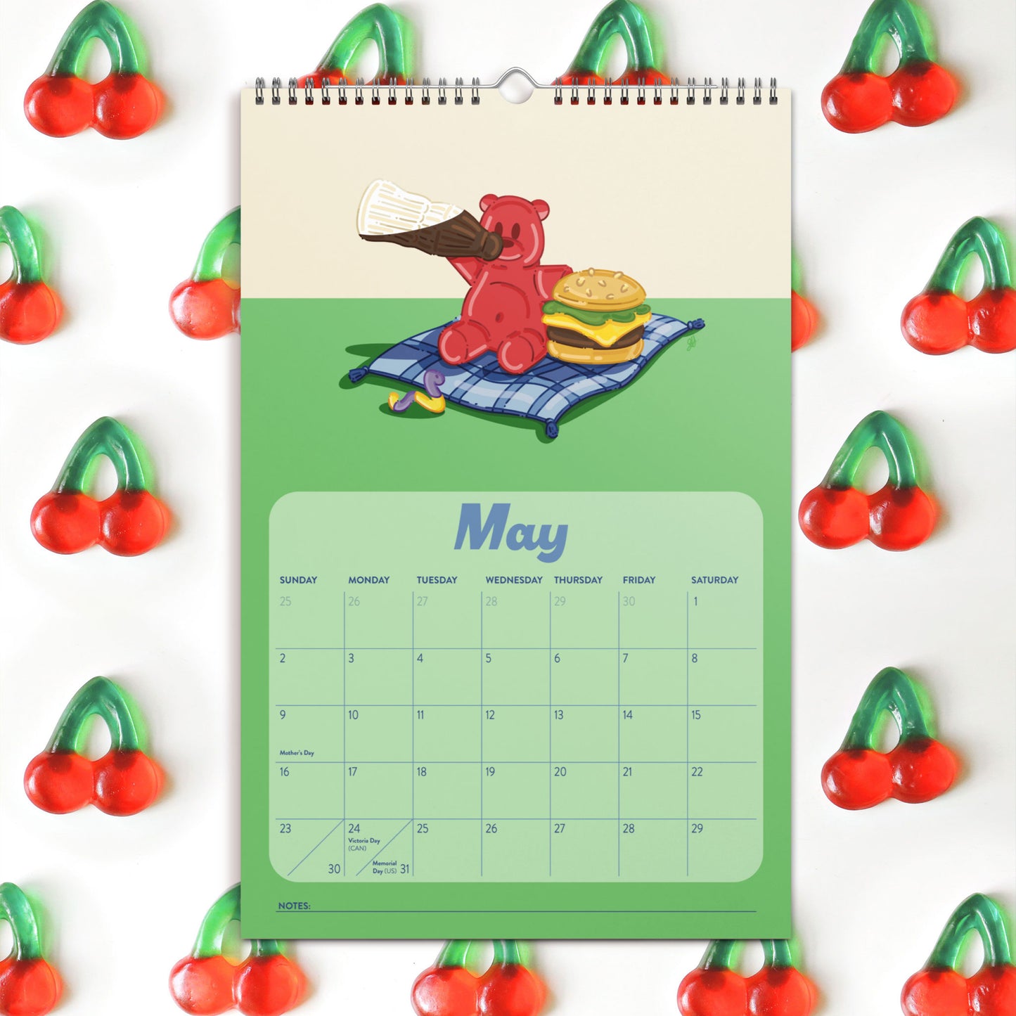 A spiral-bound monthly calendar hanging on a white background, featuring a green lower section with a May calendar grid and an illustration of a red gummy bear holding a gummy coke on a picnic blanket, with a gummy burger. The calendar is surrounded by a repeating pattern of red cherries with green stems.
