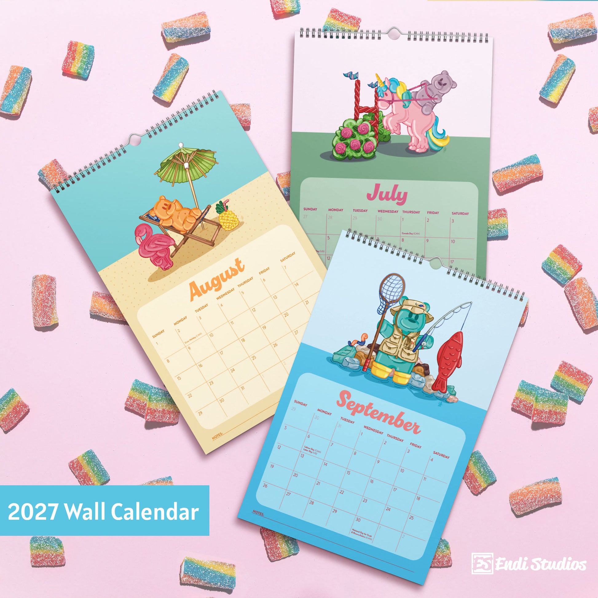 Three spiral-bound 2027 wall calendars from Endi Studios are arranged on a pink background scattered with rainbow gummy candies. The calendars feature whimsical cartoon illustrations: one shows a beach scene in August, one shows a pink pony in July, and one shows a fisherman in September. Featuring gummy bears, licorace, and swedish fish