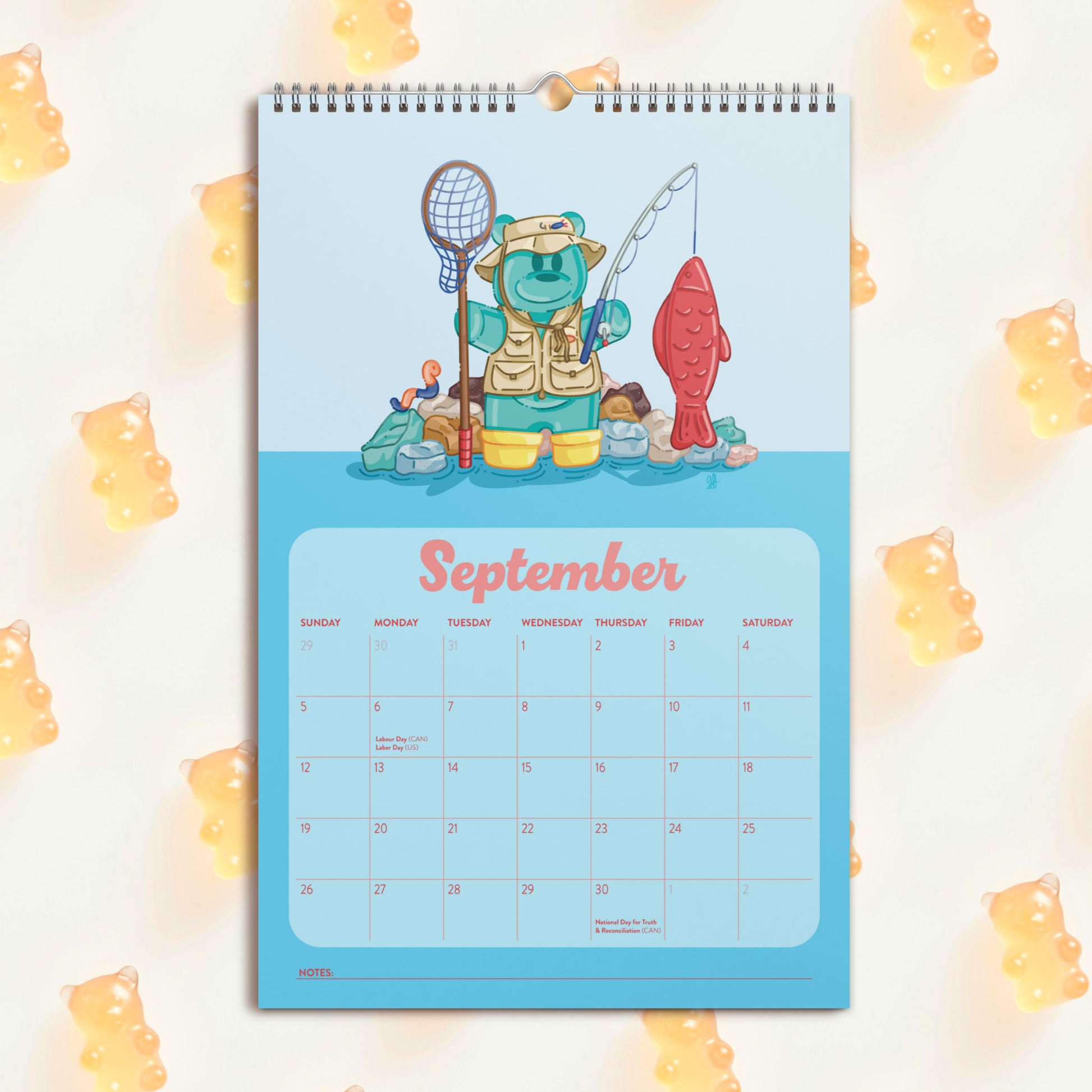 A spiral-bound September desk calendar featuring a teal gummy bear fishing with a swedish fish , set against a light background with scattered yellow gummy bear decorations.