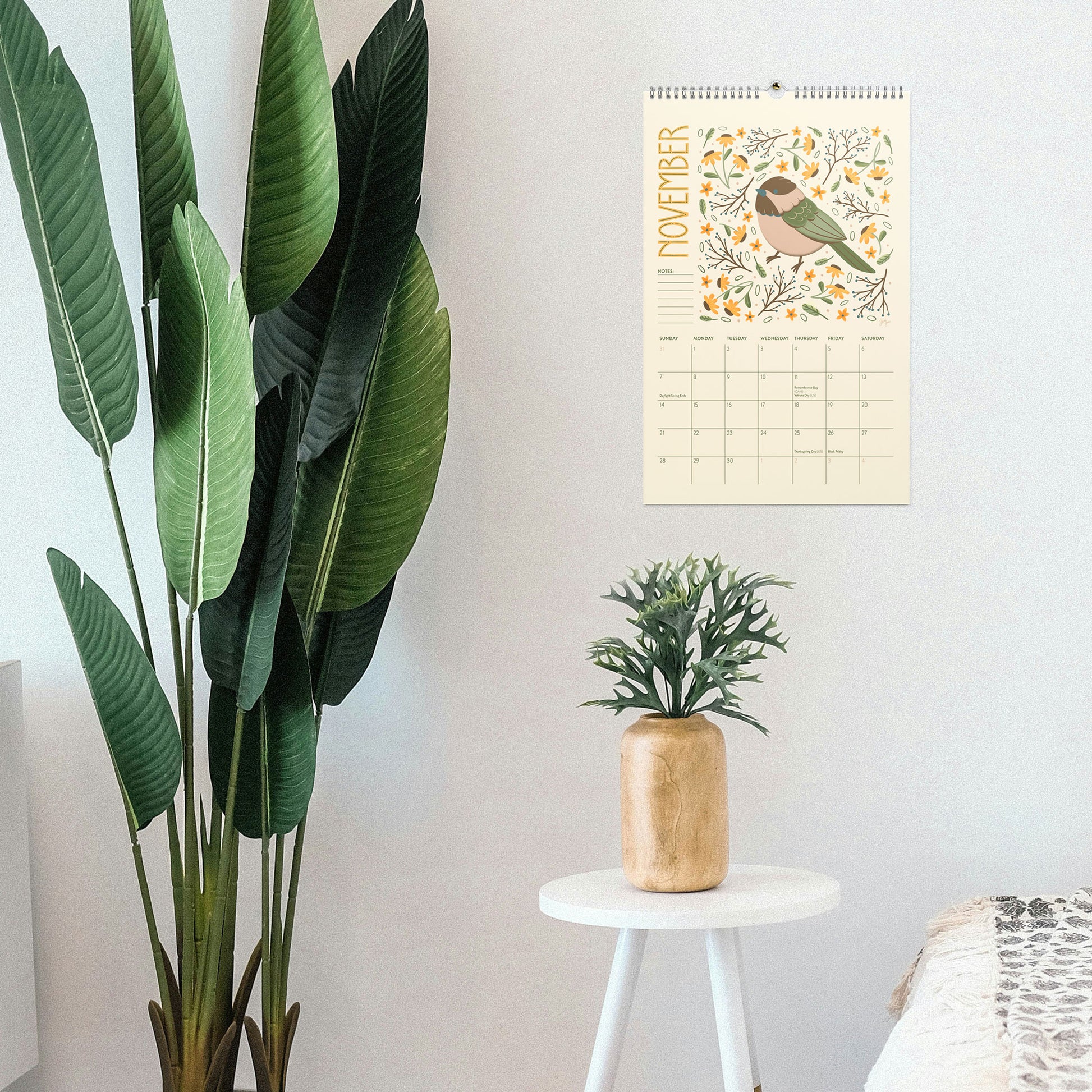 A January wall calendar featuring a bird illustration, hanging on a light gray wall in a styled room next to a large green plant and a small white side table with a gold vase. 2027 wire-bound wall calendar hanging on a neutral wall, displaying colourful folk-art animal artwork and a clear monthly layout. Ideal for home offices, kitchens, studios, classrooms, or anyone looking for a decorative illustrated 2027 wall calendar with unique art.