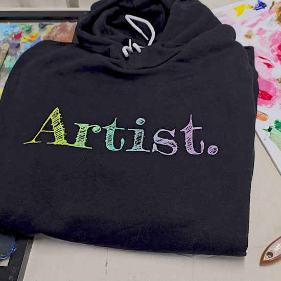 Vidoe of the Artist hoodie on an artistic background. A black artist hoodie is displayed in a flat lay. The hoodie features a sketch-style hand-drawn gradient text design in yellow, green and light purple, ideal for artists, creatives, students and illustrators. This aesthetic art hoodie is perfect for year-round outfits and makes a thoughtful gift for art lovers.