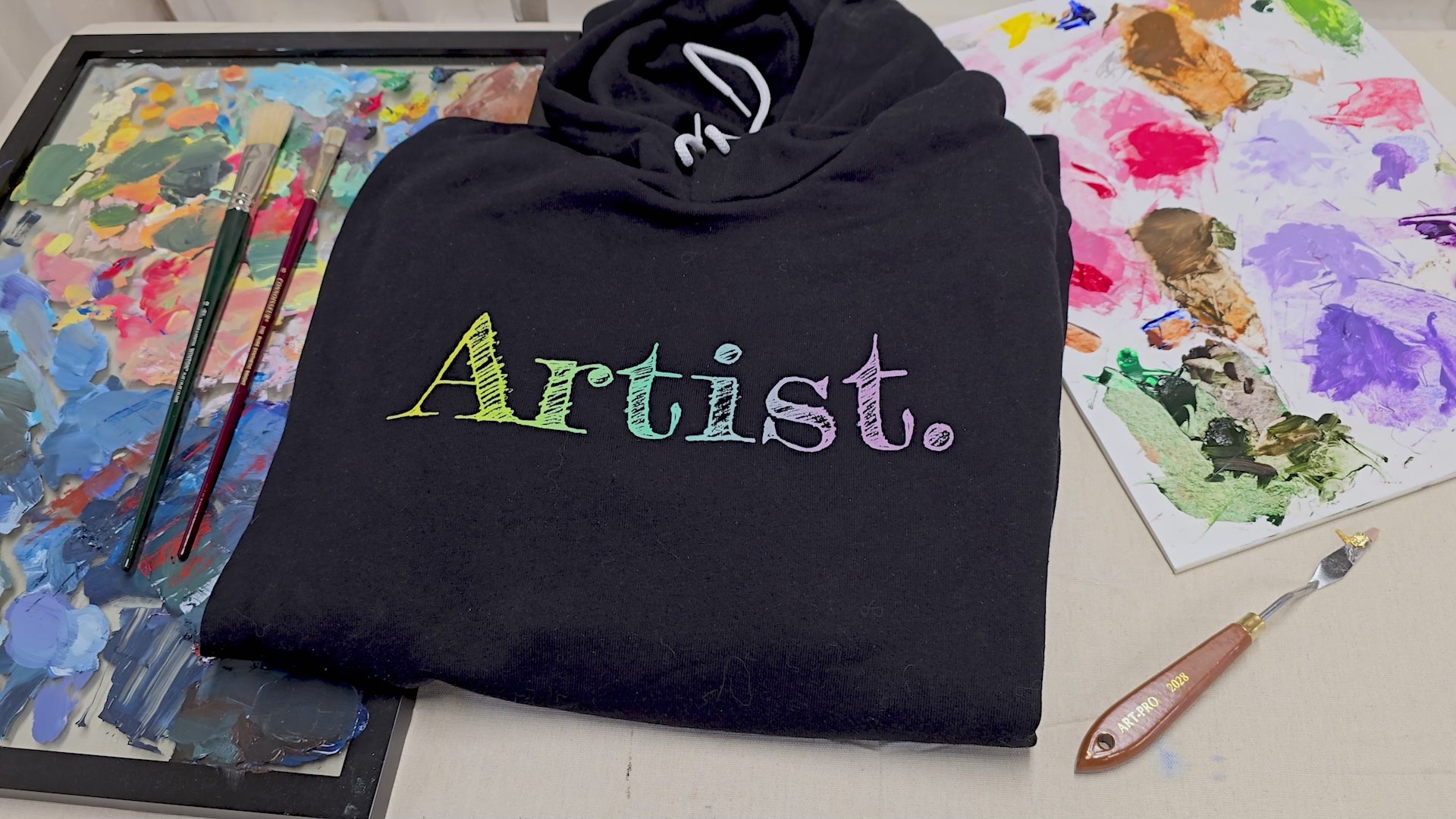 Vidoe of the Artist hoodie on an artistic background. A black artist hoodie is displayed in a flat lay. The hoodie features a sketch-style hand-drawn gradient text design in yellow, green and light purple, ideal for artists, creatives, students and illustrators. This aesthetic art hoodie is perfect for year-round outfits and makes a thoughtful gift for art lovers.